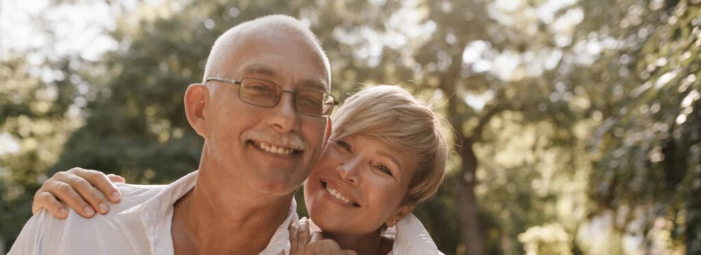 Smiling man with grey hair and mustache in glasses and light shirt hugging with blonde woman in white clothes in park.